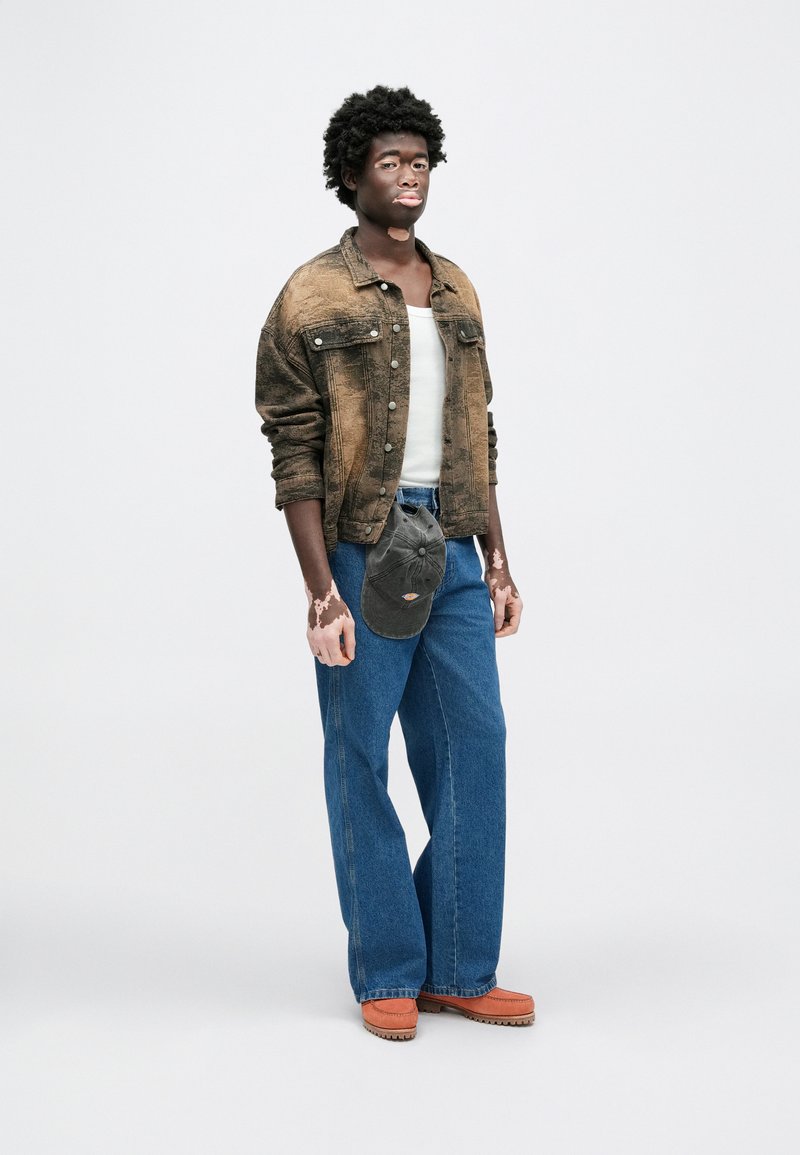 Dark brown denim jacket over a white tank top, paired with wide-leg blue jeans and orange slip-on shoes; gray cap hangs from waist.