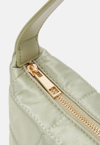 Coin d'un sac matelassé vert clair avec une sangle en cuir beige et une tirette de fermeture éclair en or, montrant des coutures détaillées et la texture.