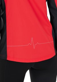 Rotes und schwarzes Sportoberteil mit weißem Herzschlagslinien-Grafik nahe der Taille, getragen mit schwarzen Leggings.