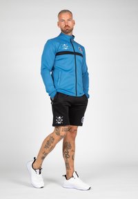 Blauwe zip-up jas met zwarte strepen, zwarte shorts met logo's, witte sneakers. Kenmerkt zichtbare tatoeages op de benen.