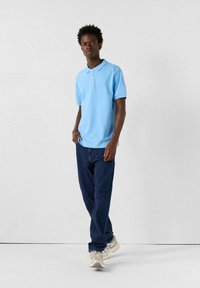 Polo shirt azzurro chiaro con maniche corte, placket con bottoni e logo ricamato; abbinato a jeans blu scuro e sneakers beige.