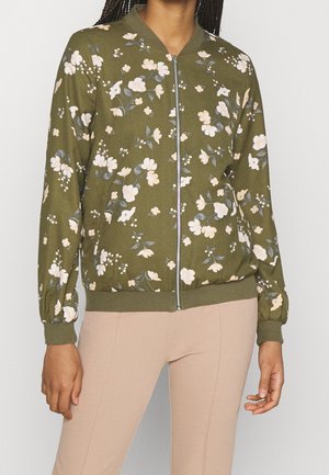 Vrouw draagt een olijfgroene jas met rits, witte bloemenprint en geribbelde manchetten, gecombineerd met lichtbeige broek.