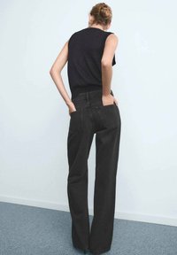 Jean large en denim noir à taille haute, avec deux poches arrière et un tissu texturé. Associé à un haut sans manches noir.