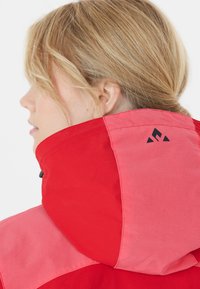 Veste à capuche rouge et rose avec un col haut, texture lisse et un détail de logo noir sur la section rose près de l'épaule.