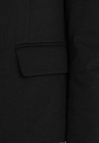 Tissu noir avec un détail de poche plate. La texture semble lisse, et le tissu est structuré avec des lignes nettes. Aucun motif ou accessoire visible.