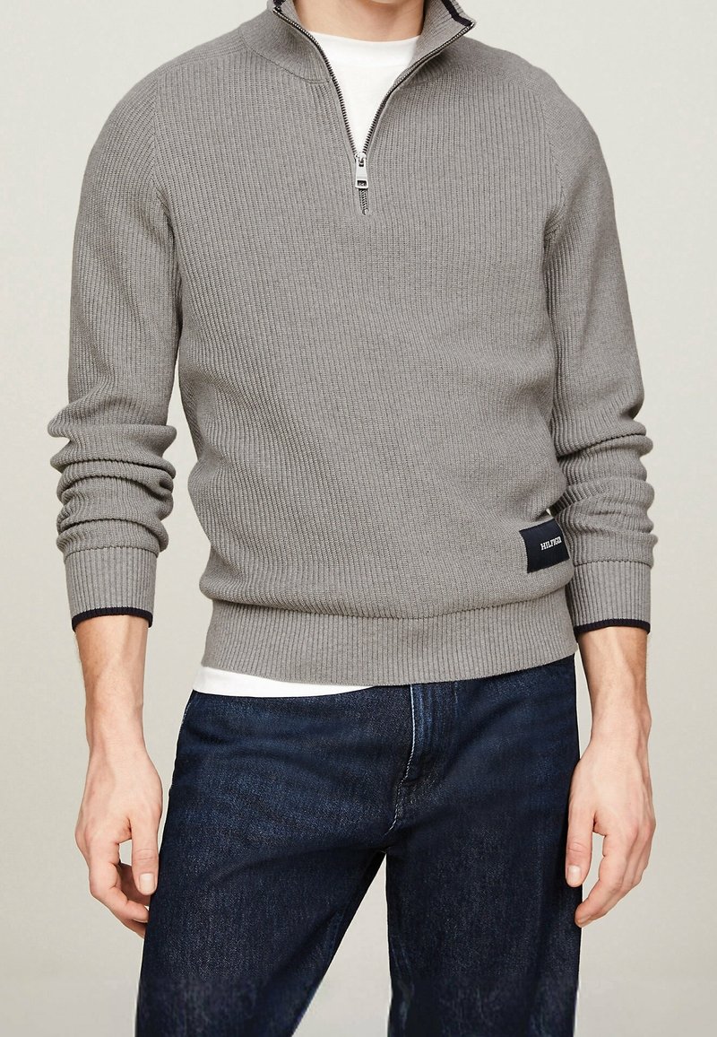 Homme portant un pull gris côtelé à col montant avec fermeture éclair sur une chemise blanche et un jean bleu foncé, debout les mains détendues le long du corps.