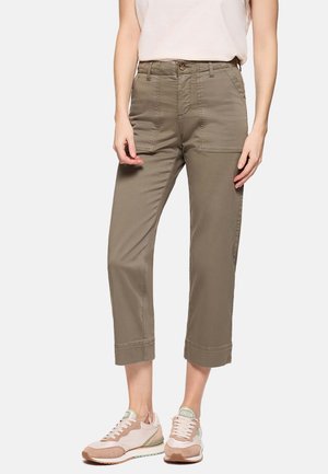 Persona vistiendo pantalones cortos de color verde oliva con bolsillos frontales de parche y zapatillas beige-rosadas, de pie contra un fondo liso.