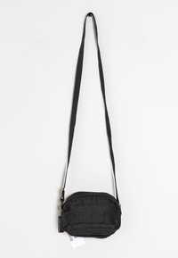 Malá černá crossbody taška se zipem, nastavitelným popruhem a visačkou na bílém pozadí.