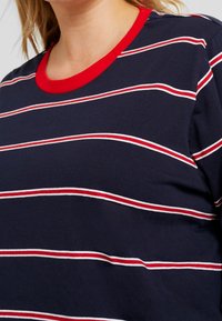 Granatowa koszulka z poziomymi czerwonymi i białymi paskami, z solidnym czerwonym ściągaczem przy szyi. Wykonana z miękkiej, elastycznej bawełny.