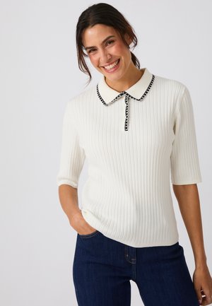 Mujer sonriente con cabello oscuro que lleva una camiseta blanca acanalada de manga corta con cuello con ribete negro y jeans azul oscuro, mano en el bolsillo.