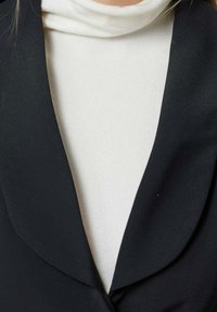 Un pull col roulé blanc porté sous un blazer noir à larges revers, avec un focus sur la zone de la poitrine et du cou.