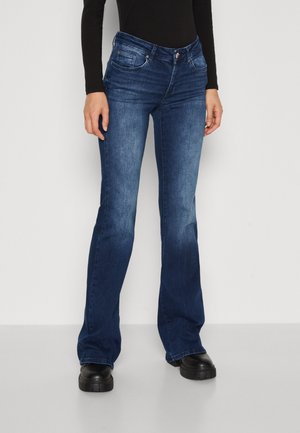 Persoon die donkerblauwe bootcut jeans, een zwart langärmelig shirt en stevige zwarte schoenen draagt, staand tegen een effen achtergrond.
