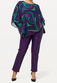 Blouse colorée et ample avec des motifs abstraits verts, violets et bleus ; associée à un pantalon en simili cuir violet ajusté et des talons noirs.