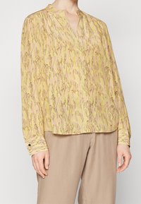 Blusa con maniche lunghe di colore giallo chiaro con motivo astratto in rosa e marrone. Presenta un profondo scollo a V e polsini con bottoni, con una leggera lucentezza.