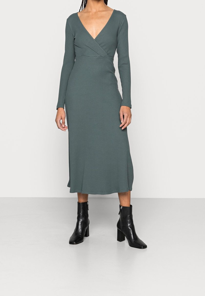 Kvinna som bär en långärmad, djup V-ringad, ribbad grön midiklänning med svarta klackiga ankelskottar, stående mot en ljusgrå, enfärgad bakgrund.