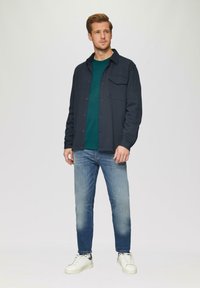 Marineblauwe overhemd met knopen over een donkergroene ronde hals, gecombineerd met lichtblauwe jeans en witte sneakers met grijze accenten. Gladde stof, aansluitend ontwerp.