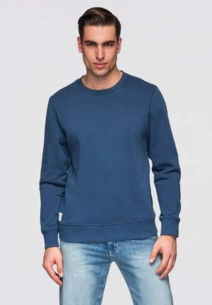 Nuori mies, jolla on taaksepäin kammatut hiukset, pukeutunut yksiväriseen siniseen collegepaitaan ja vaaleansinisiin farkkuihin, seisoo valkoista taustaa vasten.