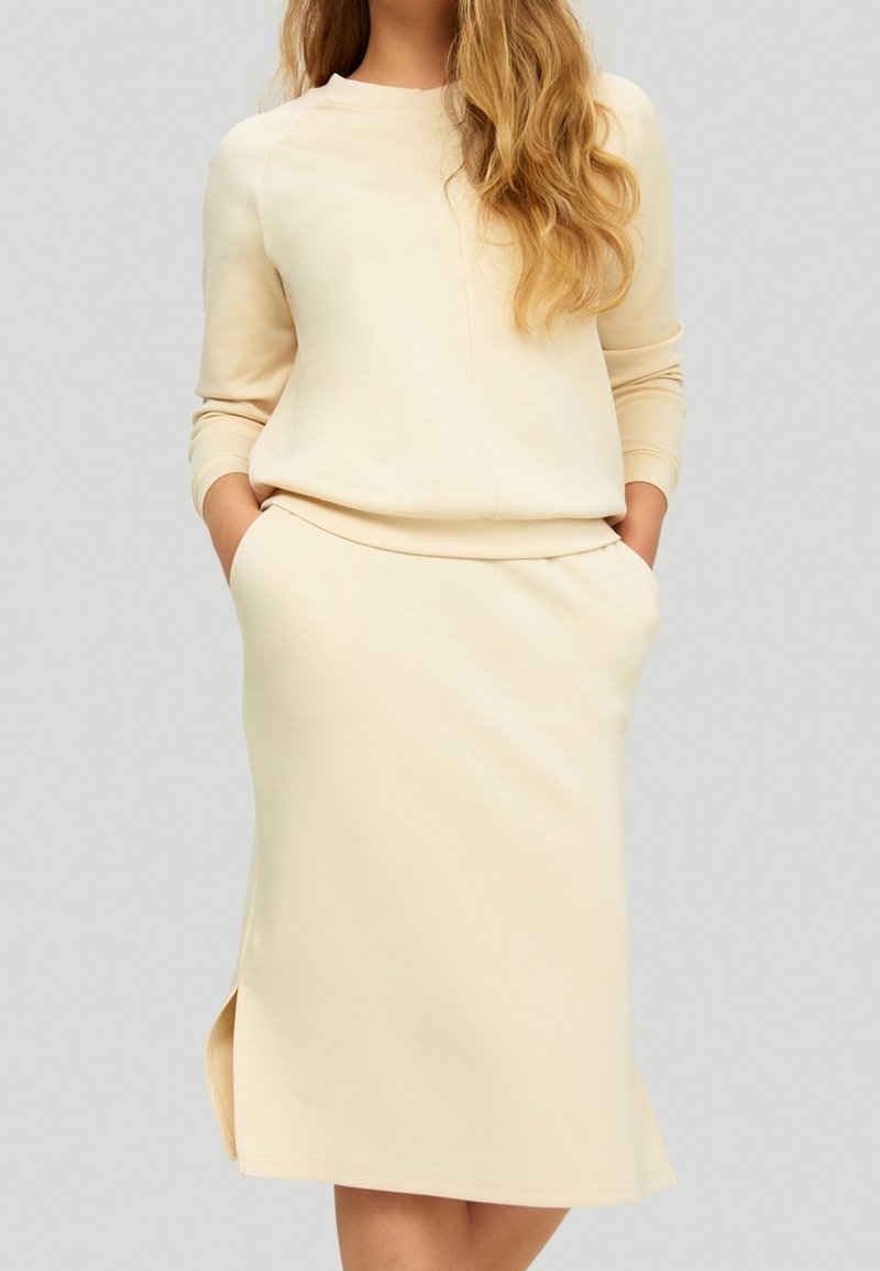Femme portant un pull crème à manches longues et une jupe assortie longueur genou avec poches et fente latérale, sur un fond gris clair uni.