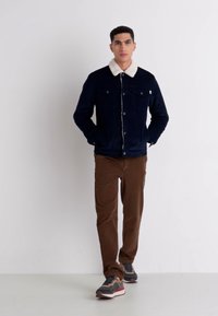 Jachetă din denim bleumarin cu guler din blană sintetică albă, buzunare frontale, asortată cu pantaloni din corduroy maro și adidași gri. Stil casual.