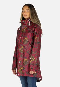 Kvinde iført en rød regnjakke med blomstermønster, høj krave og hvide snore, kombineret med blå jeans.