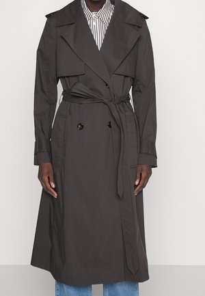 Personne portant un trench-coat noir ceinturé par-dessus une chemise rayée à boutons et un jean bleu, debout devant un fond gris uni.