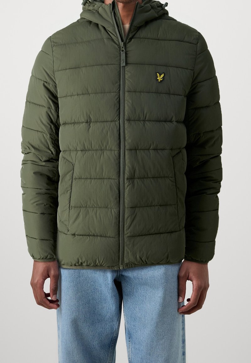Homme portant une veste matelassée olive à fermeture éclair avec capuche et logo d'oiseau jaune sur la poitrine, associée à un jean bleu clair.