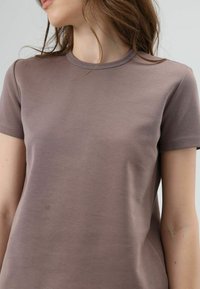 Bruine T-shirt met korte mouwen en een ronde hals. Gemaakt van zachte, gladde stof. Heeft subtiele stiksel-details langs de naden.