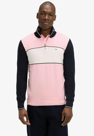 Homme portant un pull-over à manches longues rose, blanc et bleu marine avec un col à zip quart, et un pantalon foncé, debout devant un fond uni.