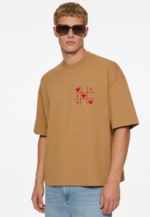 Junger Mann mit Schildpatt-Sonnenbrille und einem beigen, übergroßen T-Shirt mit rotem Tic-Tac-Toe-Design mit Herzen und Kreuzen auf der Brust.