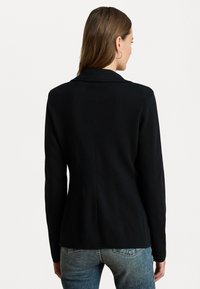 Zwart blazer met een gestructureerd ontwerp, slim fit en een opstaande kraag, met een gladde textuur en minimale details. Gemaakt in combinatie met blauwe jeans.