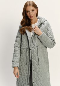 Grau gesteppter langer Mantel mit Kapuze, Kordelzug in der Taille, seitlichen Taschen und Druckknopfverschlüssen, mit glatter, glänzender Textur.