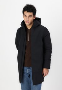Svart hettejakke med glidelås foran, elegant design og quiltet tekstur. Båret over en brun genser og blå jeans.