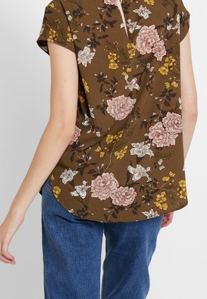 Vue de dos d'une personne portant un chemisier marron à fleurs à manches courtes avec des fleurs roses, blanches et jaunes et un jean bleu.