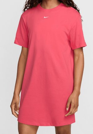 Femme portant une robe t-shirt Nike corail à manches courtes avec un logo Nike blanc sur la poitrine, debout devant un fond uni.