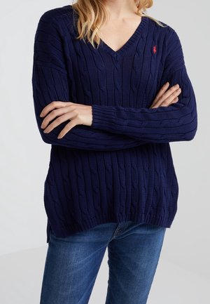 Kvinde iført en marineblå V-hals kabelstrikket sweater med korslagte arme og blå jeans foran en ensfarvet baggrund.