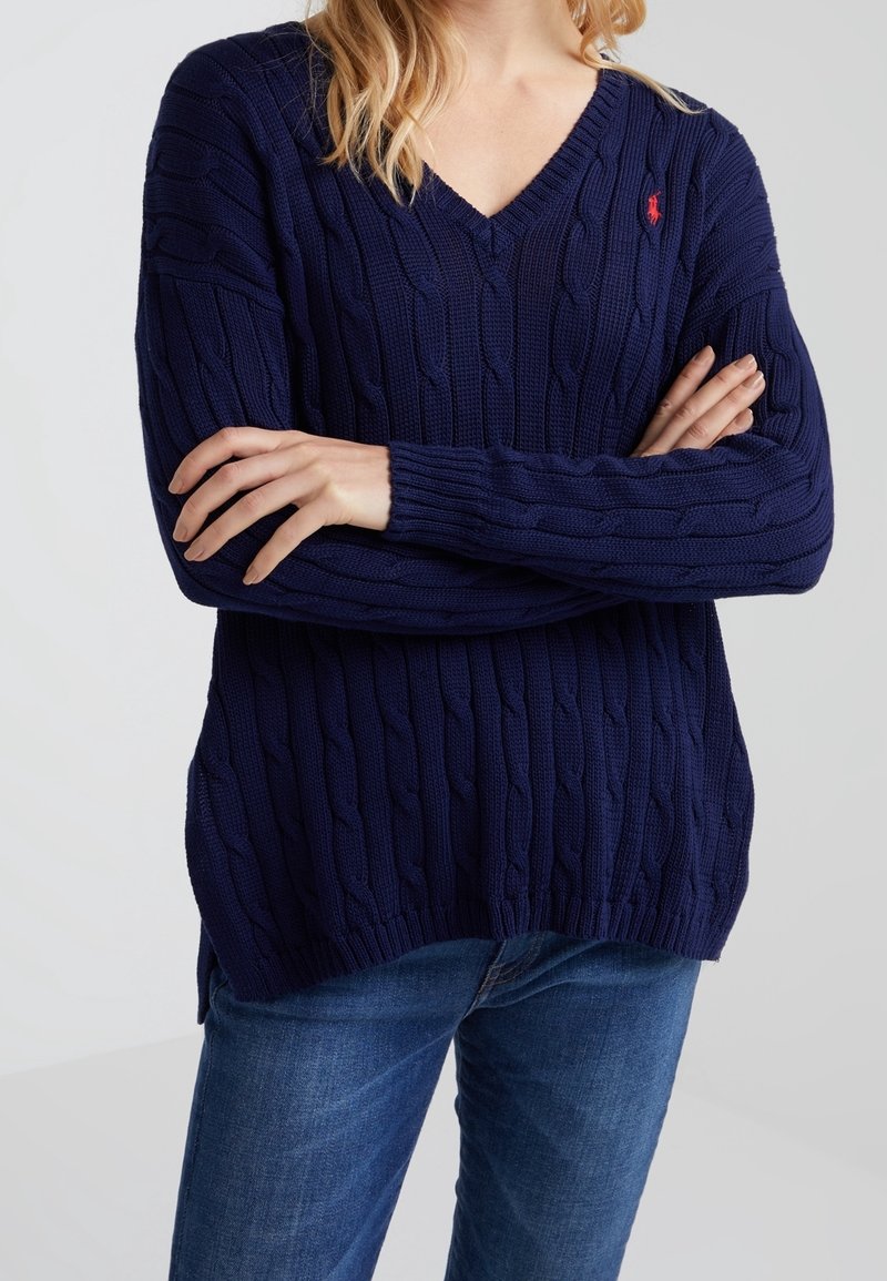 Femme portant un pull à col en V en maille torsadée bleu marine, les bras croisés, et un jean bleu, devant un fond uni.