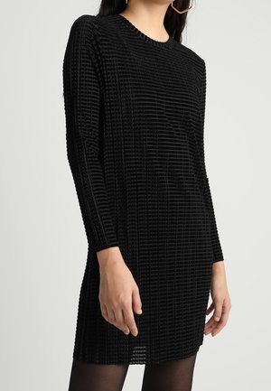Frau trägt ein langärmeliges, schwarzes, texturiertes Mini-Kleid mit horizontalem Rippenmuster und schwarze transparente Strumpfhosen, steht vor einem einfarbigen Hintergrund.