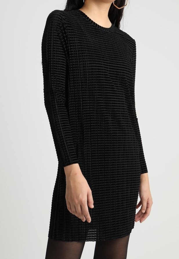 Femme portant une mini-robe noire texturée à manches longues avec un motif côtelé horizontal et des collants noirs transparents, debout devant un fond uni.