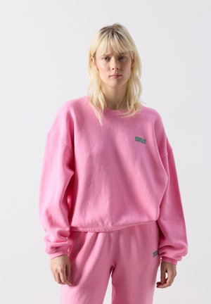 Junge Frau mit hellen Haaren, die einen passenden pinkfarbenen Pullover und Jogginghose mit kleinen grünen "American Vintage"-Logos trägt.