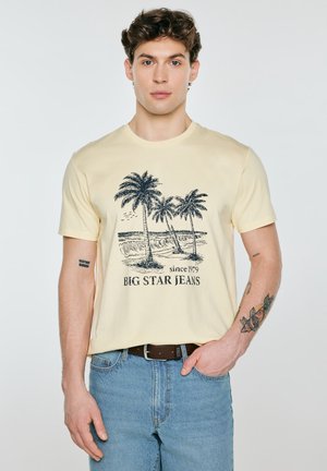 Mladý muž s krátkými kudrnatými vlasy, oblečený v béžovém tričku s potiskem palmy a modrých džínách, stojící před jednobarevným pozadím.