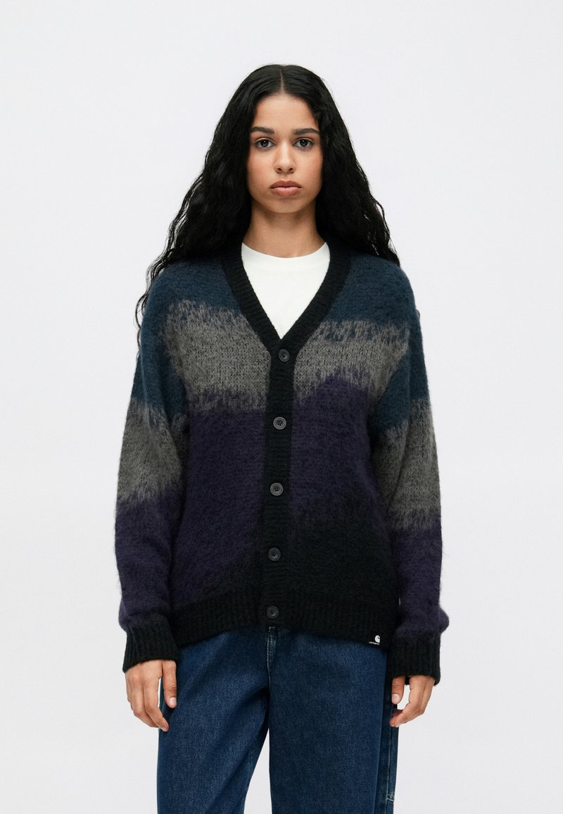 Woman with long dark hair wearing a buttoned, fuzzy cardigan with blue, gray, and purple stripes over a white shirt and loose blue jeans.