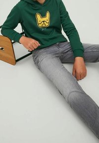 Groene hoodie met een gele hondenafbeelding op de voorkant, gecombineerd met vervaagde grijze jeans. De hoodie heeft ribgebreide manchetten en een voorkantzak.