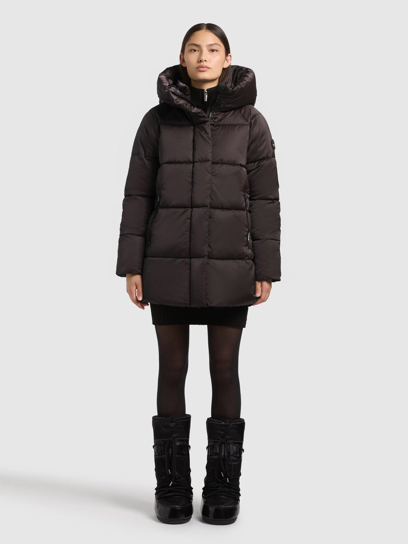 Svart puffjacka med quiltat mönster och hög krage. Har en stor huva, dragkedja framtill och sidfickor. Bärs med en svart kjol och stövlar.