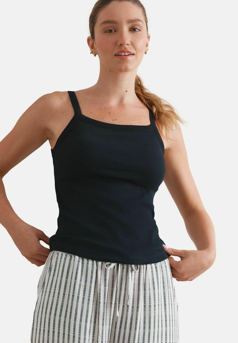 Femme à la peau et aux cheveux clairs, avec une queue de cheval, portant un haut sans manches noir et un pantalon blanc avec des rayures verticales noires, en train d'ajuster son haut.