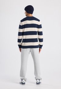 Uomo che indossa un maglione a righe blu navy e crema, pantaloni della tuta grigi, berretto a maglia blu navy e sneaker bianche, in piedi di spalle davanti a uno sfondo semplice.