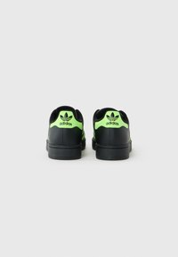 Une paire de baskets Adidas noires avec des onglets de talon vert fluo, affichant le logo Adidas, photographiée de dos sur un fond uni.