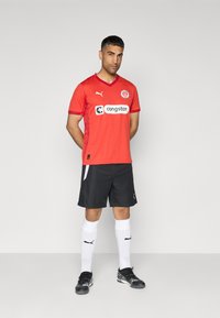 Maillot de football rouge à manches courtes, avec un logo blanc et un motif sur les côtés, associé à un short noir, des chaussettes blanches et des crampons noirs.