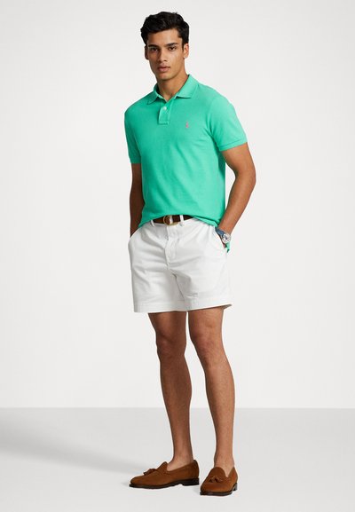 Polo vert, shorts blancs, mocassins bruns. Le polo présente un col, des manches courtes et un petit logo brodé sur la poitrine.
