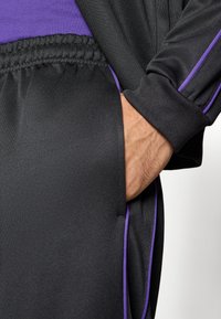 Μαύρο αθλητικό σύνολο με μοβ λεπτομέρειες. Διαθέτει υφή υφάσματος, ελαστική μέση και χέρι  τοποθετημένο στην τσέπη.