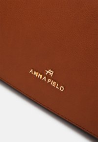 Ruskea nahkainen käsilaukku, jossa on sileä pinta, kullalla painettu logo ja ommellut reunat rakenteen takaamiseksi. Kompakti ja tyylikäs muotoilu.